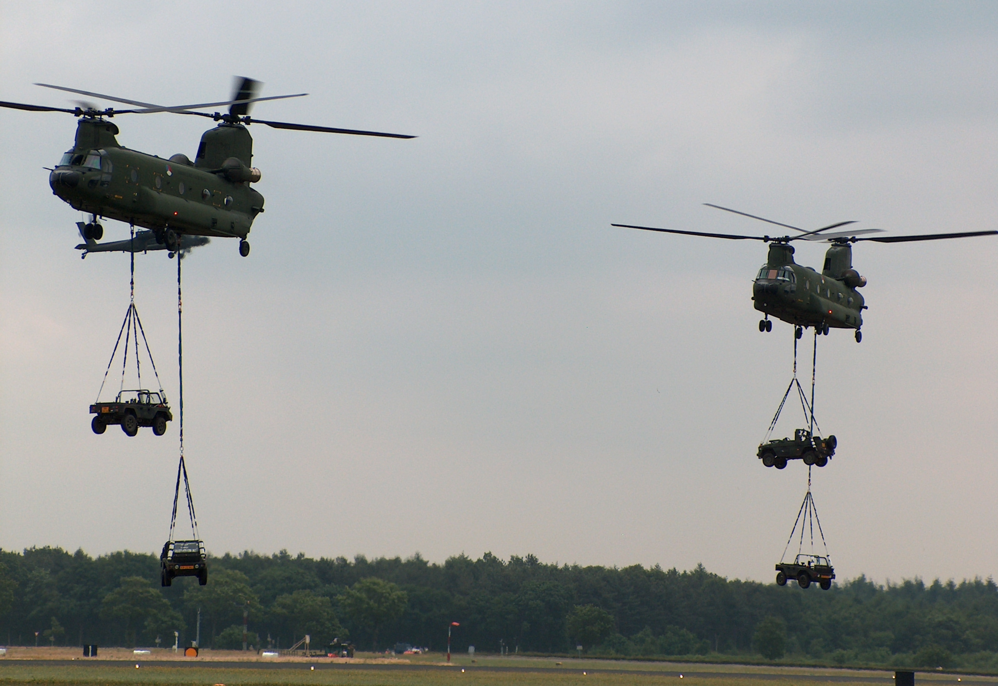 Two Chinook helicopters transporting defence land vehicles across a field